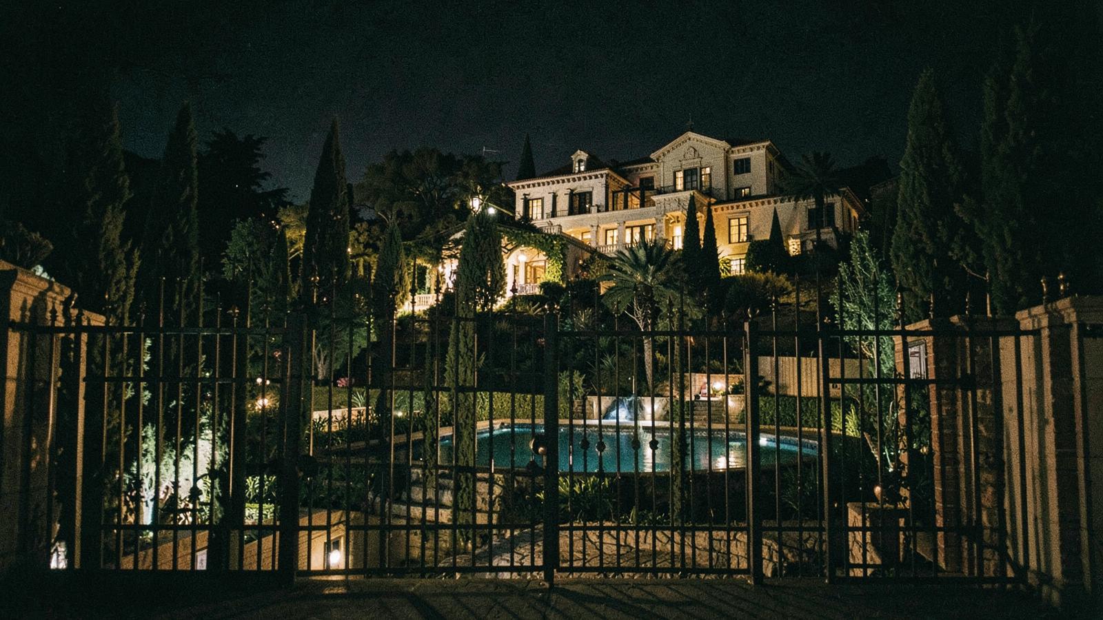 A guarded hillside estate seen through iron gates.